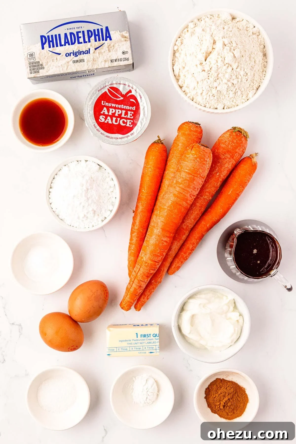 Various ingredients laid out on a kitchen counter, including flour, cinnamon, applesauce, grated carrots, and cream cheese.