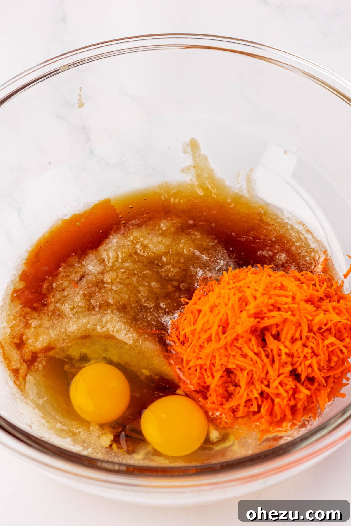 A bowl filled with wet ingredients: applesauce, grated carrots, melted butter, eggs, vanilla extract, and maple syrup.