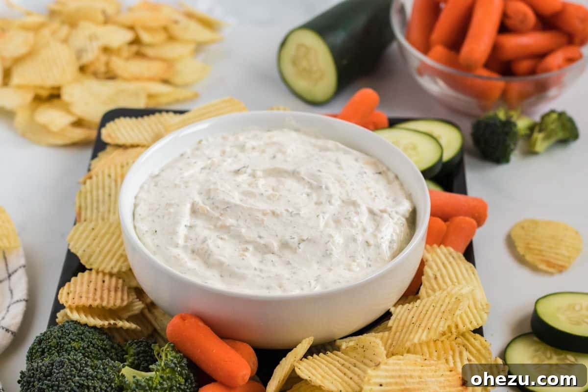 A bowl of creamy homemade chip dip, encircled by a colorful selection of dippers including potato chips, broccoli florets, baby carrots, and cucumber slices.