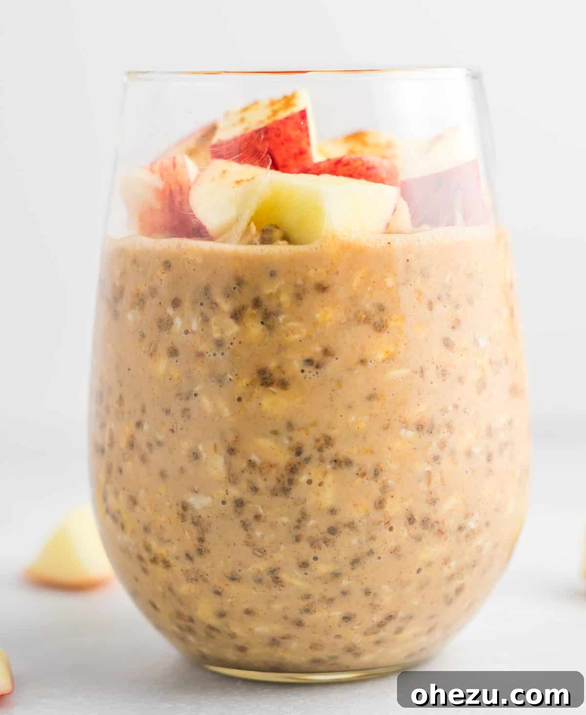 Close-up of overnight oats in a clear glass jar, showing the layers of oats, milk, and chia seeds, with a few apple chunks visible and cinnamon sprinkled on top.
