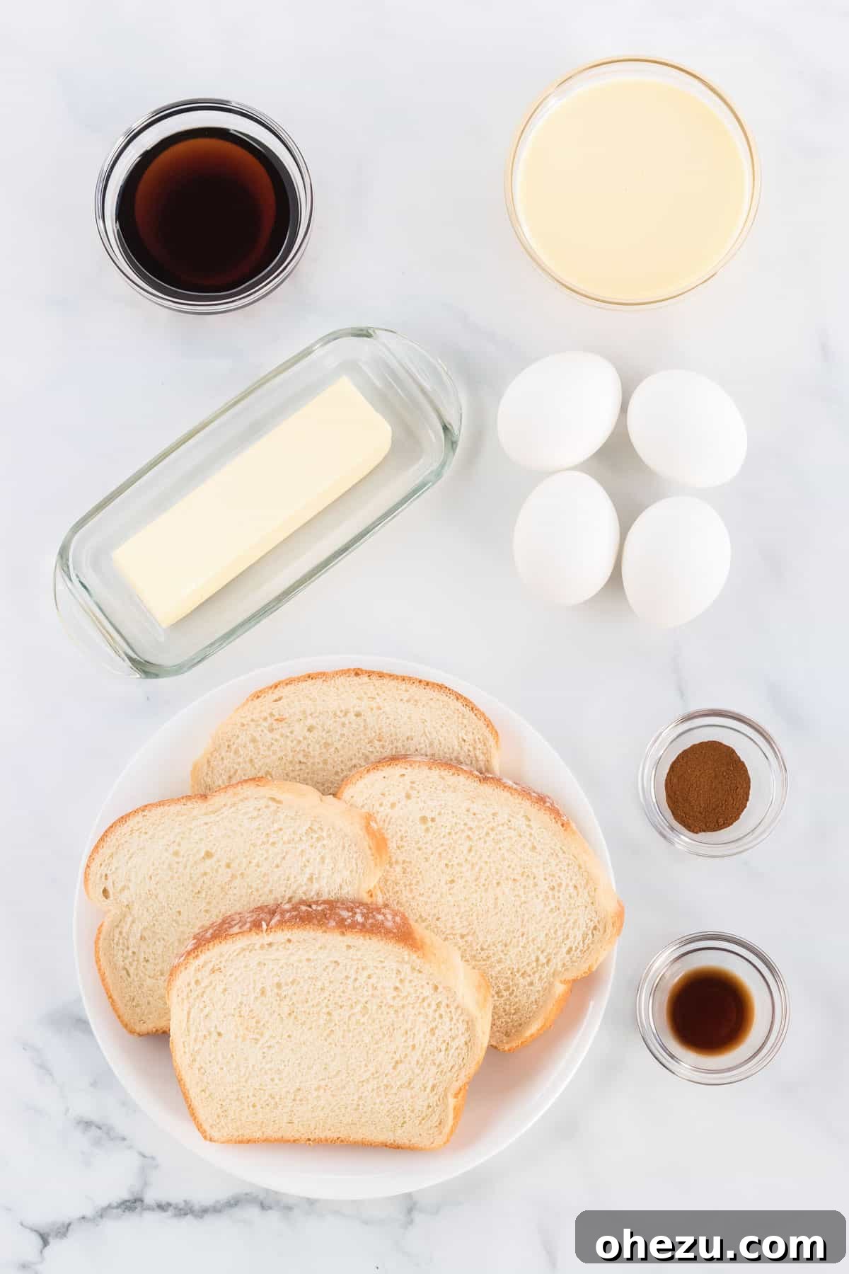 Festive Eggnog French Toast 3 A flat white dish containing all the raw ingredients needed to make eggnog French toast: a bowl of beaten eggs, a carton of eggnog, a sprinkle of cinnamon, a bottle of vanilla extract, a loaf of bread, and a bottle of rich maple syrup.