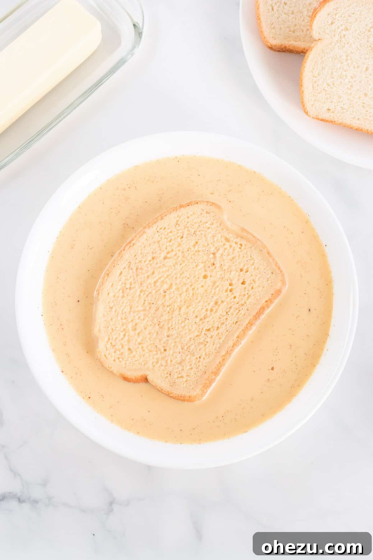 Festive Eggnog French Toast 4 A hand carefully dipping a thick slice of white bread into a shallow dish filled with the creamy eggnog French toast batter, ensuring an even and thorough coating on both sides.