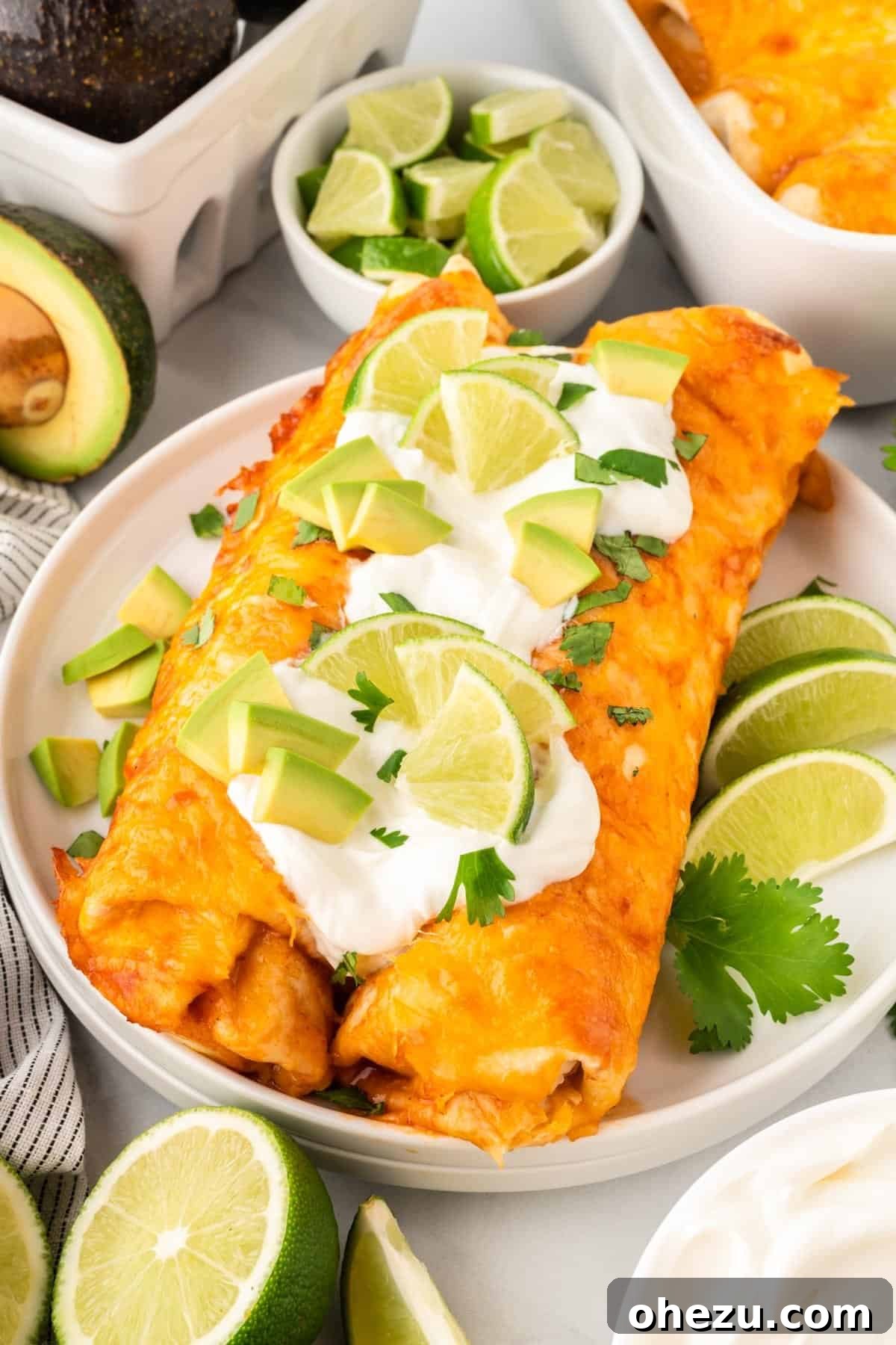 A serving of two sweet potato black bean enchiladas on a plate, garnished with fresh sour cream, ripe avocado slices, and a lime wedge.