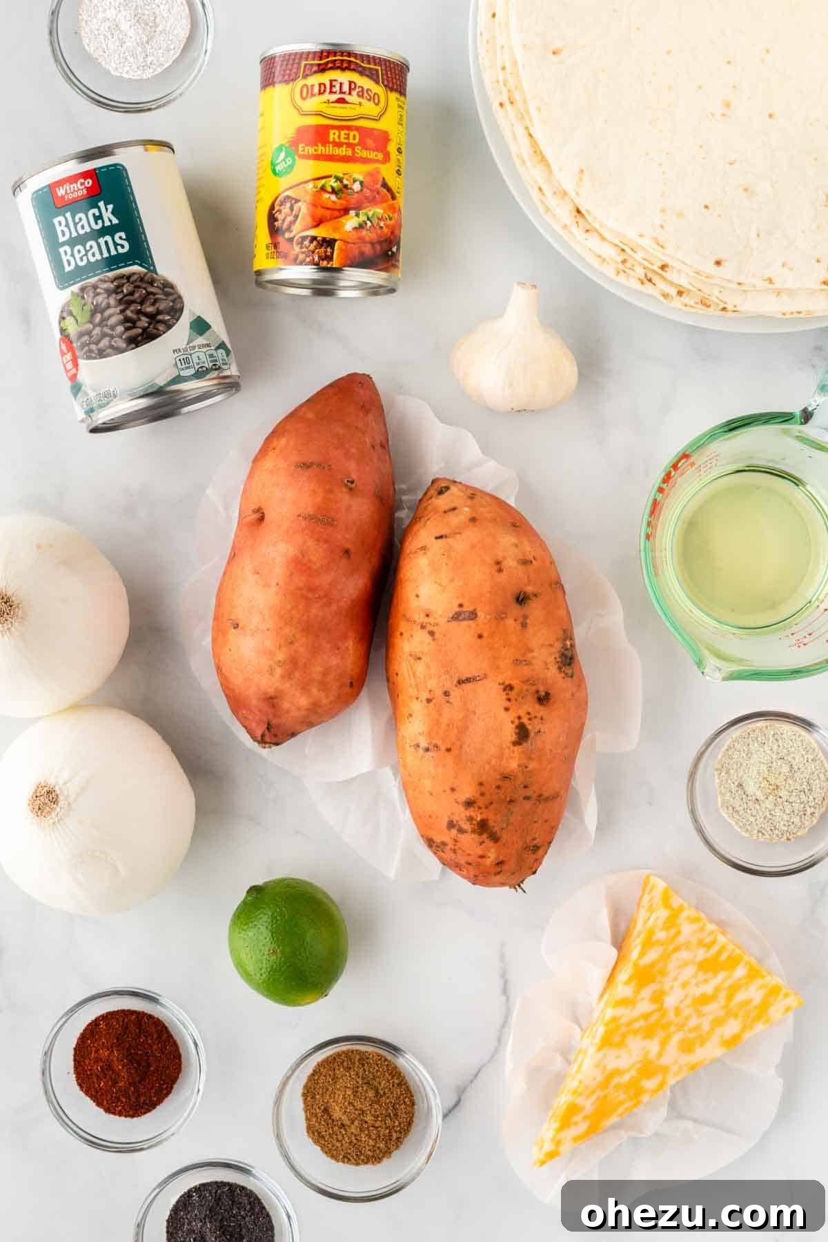 A flat lay photograph displaying all the fresh ingredients needed to make sweet potato black bean enchiladas: sweet potatoes, black beans, lime, onion, garlic, spices, cheese, enchilada sauce, and tortillas.
