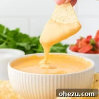 A tortilla chip being dipped into a bowl of homemade nacho cheese sauce.