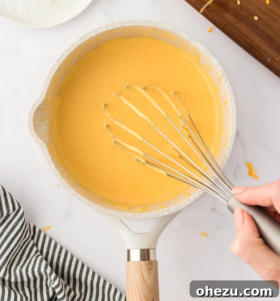5-Minute Creamy Nacho Cheese Dip 7 A metal whisk vigorously stirring a creamy, golden cheese sauce in a saucepan on the stove, showing the thickening process.