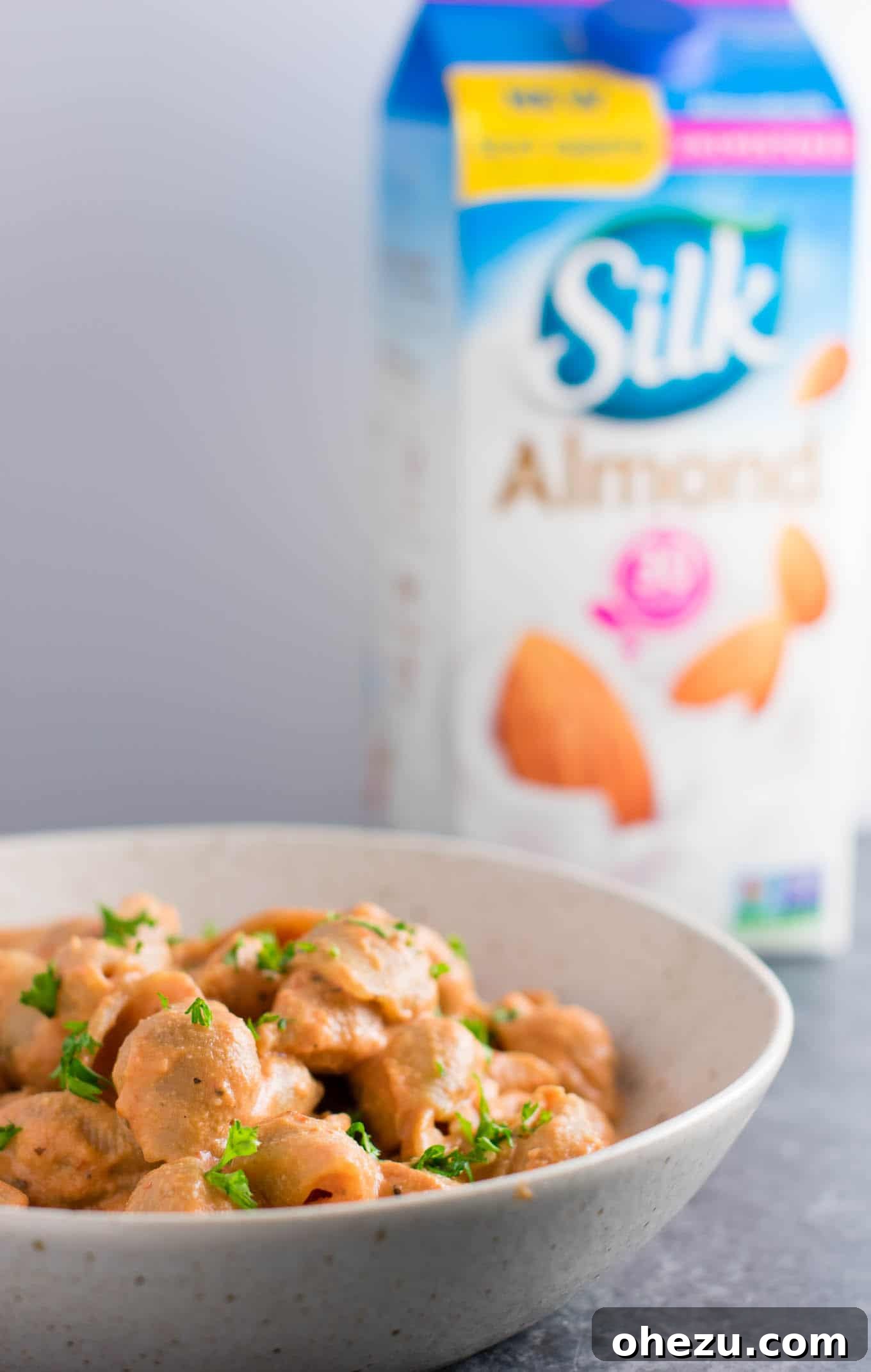 A hand pouring Silk Original Almond Milk into a measuring cup, with creamy tomato shells in the background, illustrating the key ingredient.