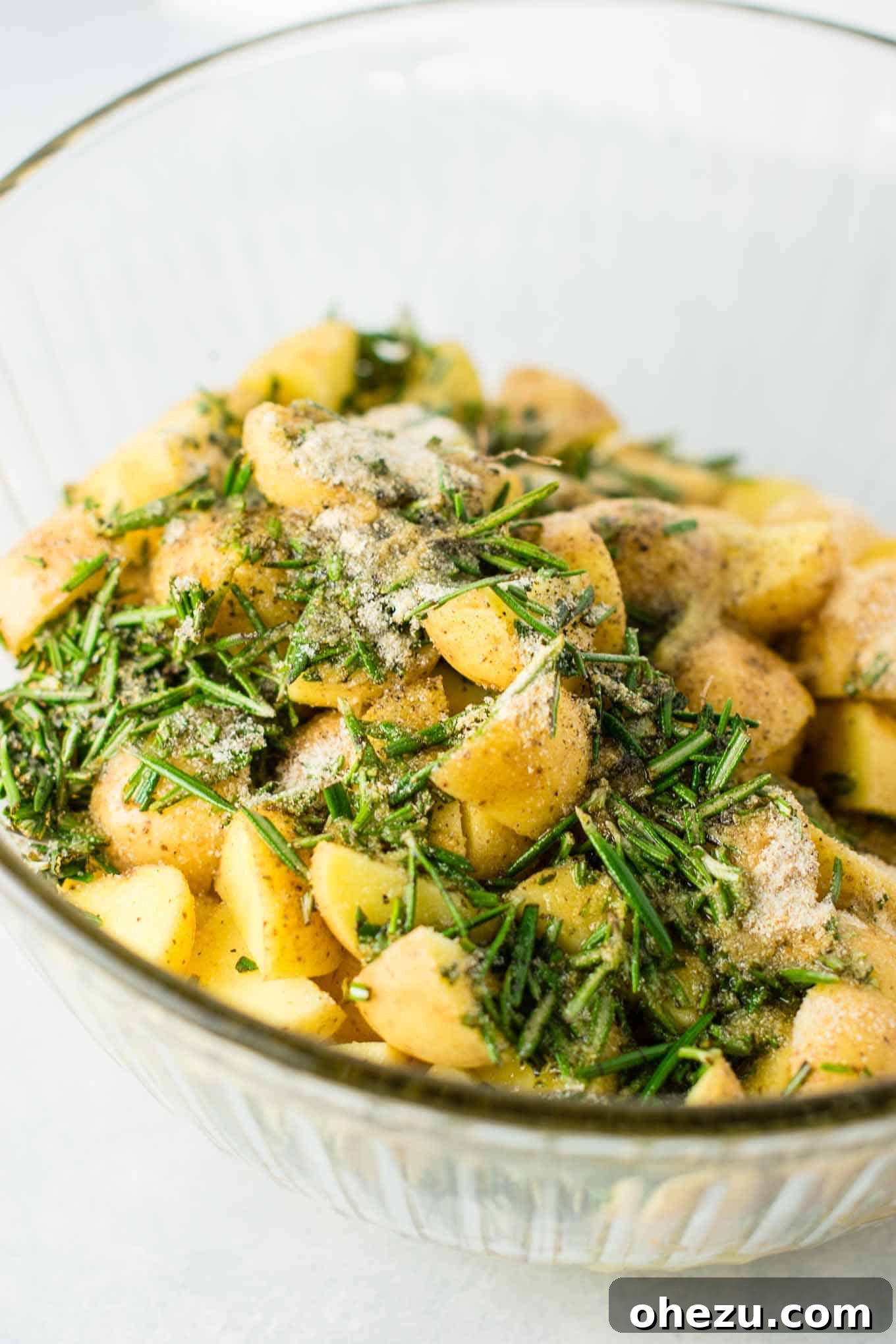A hand drizzling olive oil over the seasoned, chopped potatoes and fresh rosemary in a bowl, ensuring an even coat.