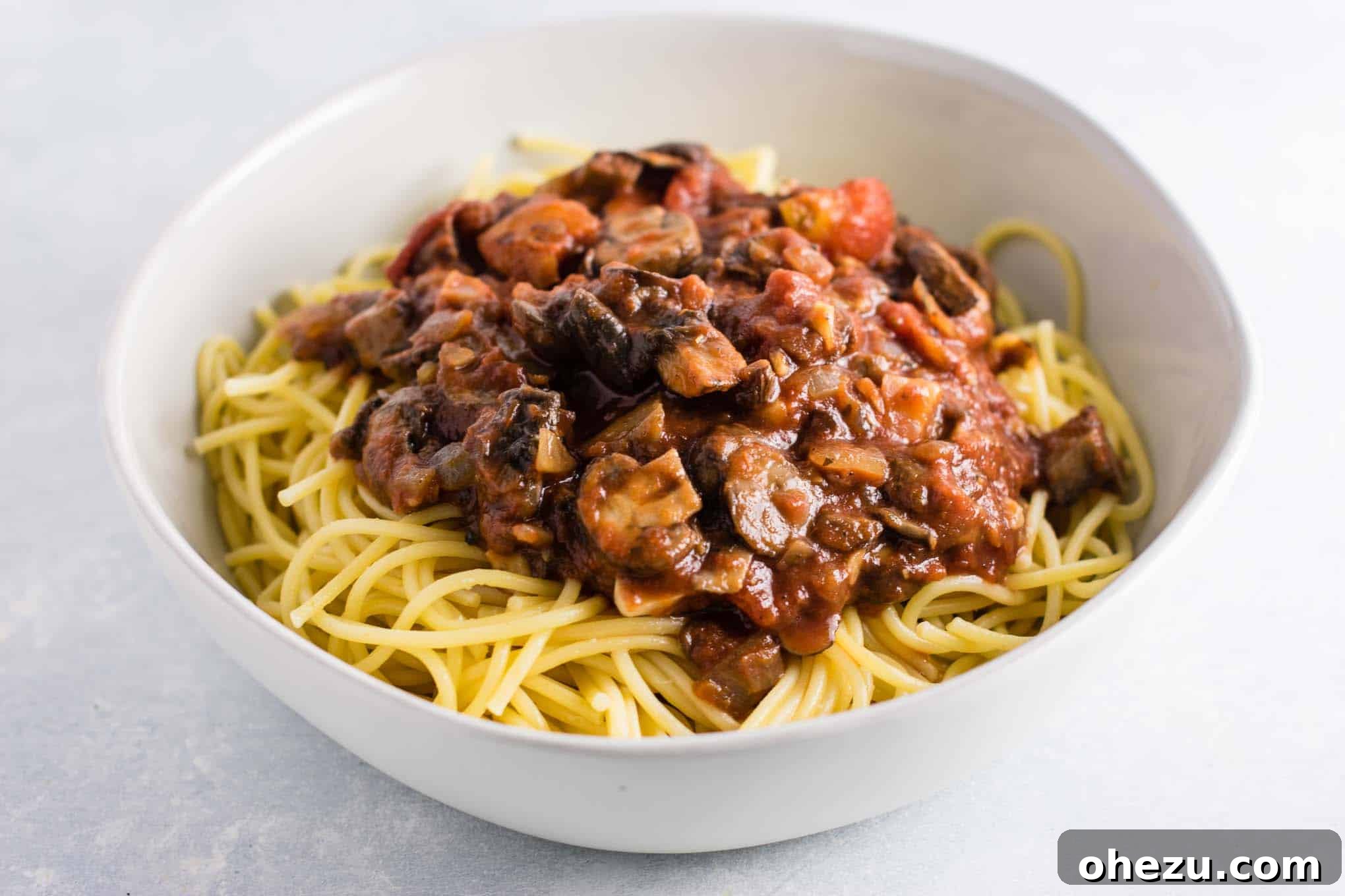 A generous serving of hearty vegetarian spaghetti sauce, rich with mushrooms and aromatics, beautifully plated over tender white spaghetti noodles in a deep white bowl.