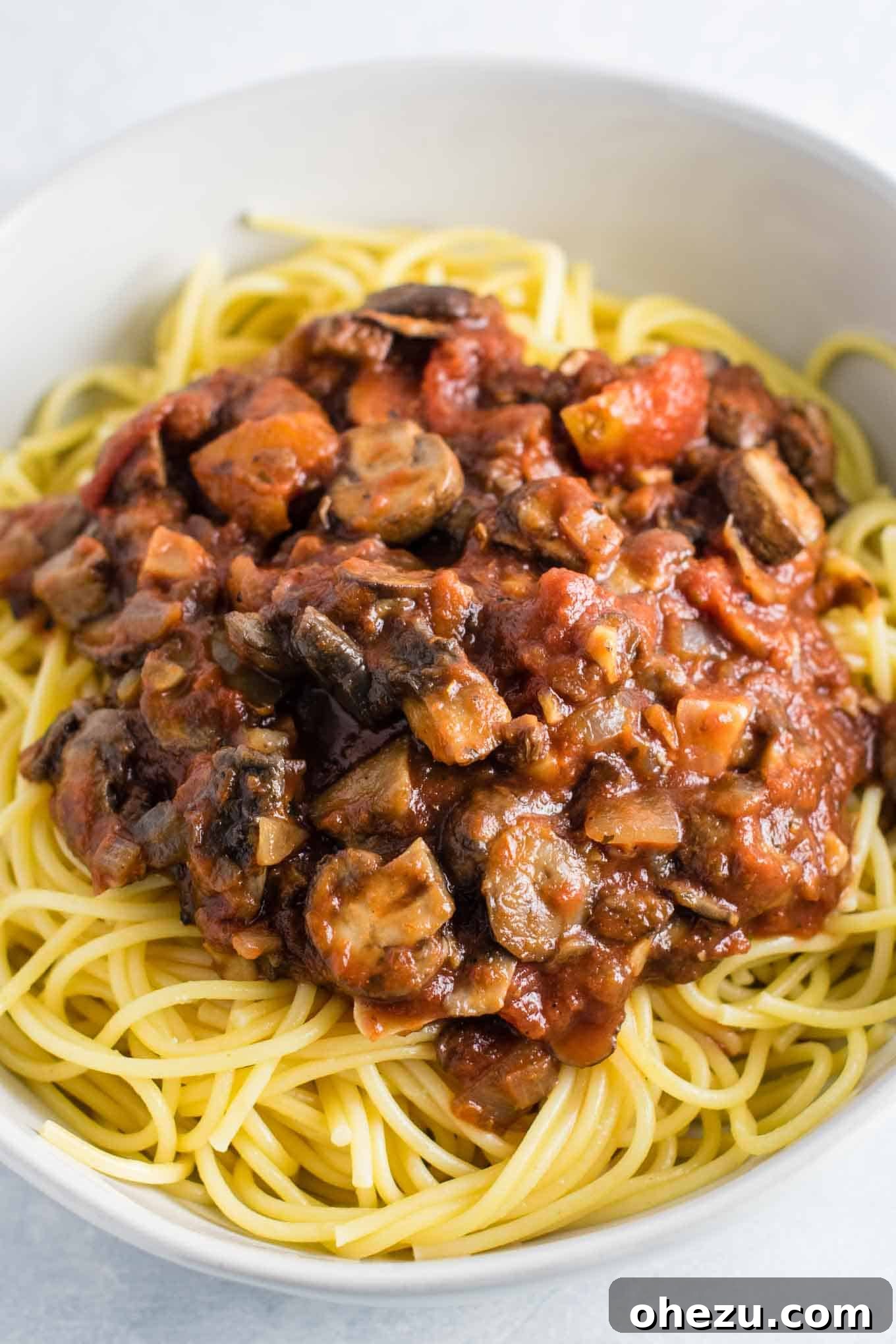 Close-up of chunky mushroom pasta sauce richly coating spaghetti noodles in a white bowl, highlighting the texture of the vegetables.