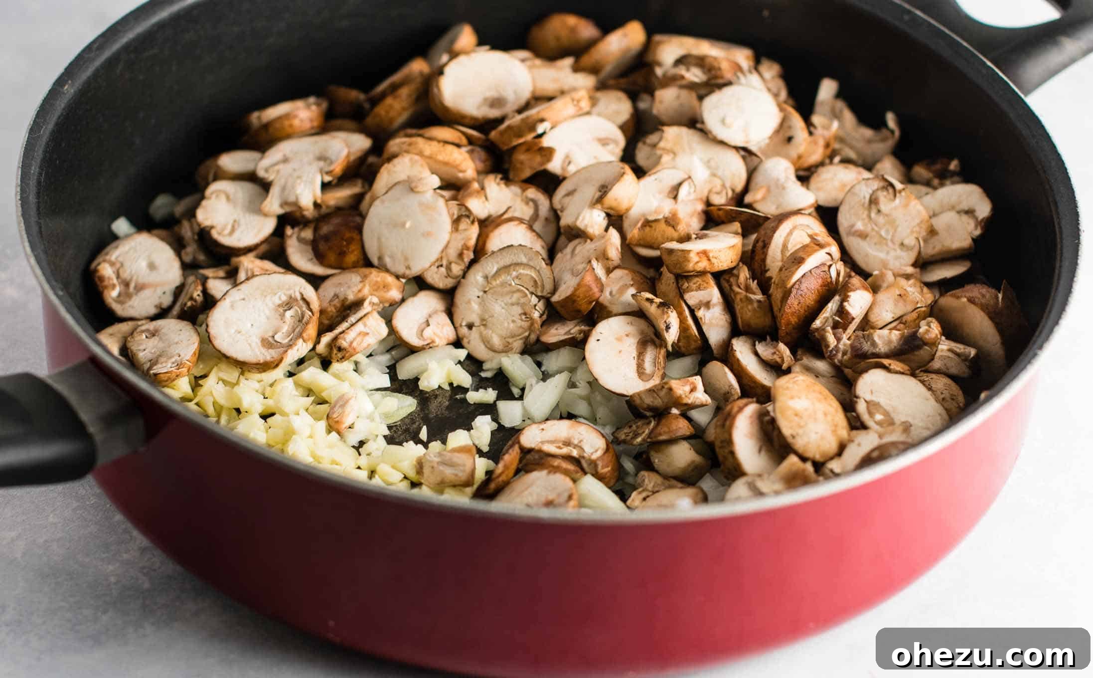 A vibrant mix of sliced baby bella mushrooms, diced yellow onion, and minced garlic simmering gently in a large, bright red skillet, forming the savory base of the pasta sauce.