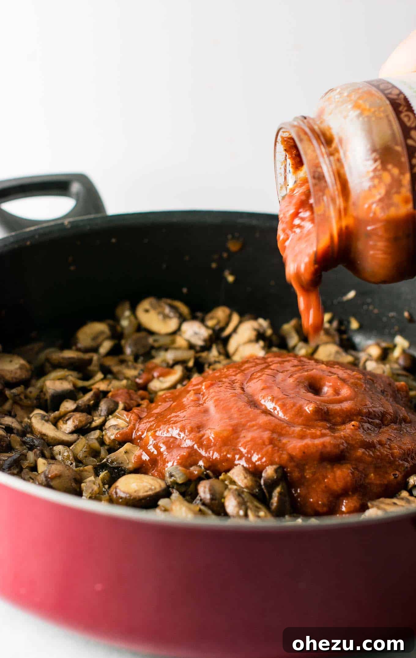 A rich, red pasta sauce being poured directly into the skillet, coating the beautifully cooked mushroom, garlic, and onion mixture.