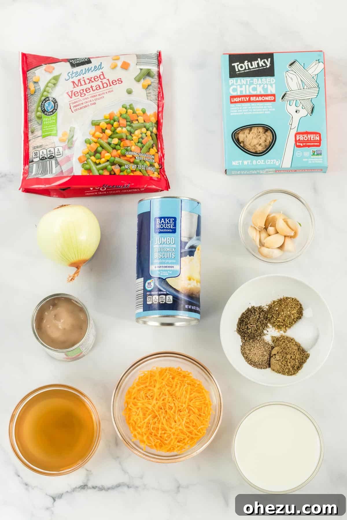 A flat lay photograph showcasing all the fresh and frozen ingredients needed to prepare a delicious vegetarian pot pie, including onions, garlic, frozen mixed vegetables, cream of mushroom soup, vegetable broth, milk, sharp cheddar cheese, various herbs, meatless chick'n strips, and ready-to-bake biscuits.