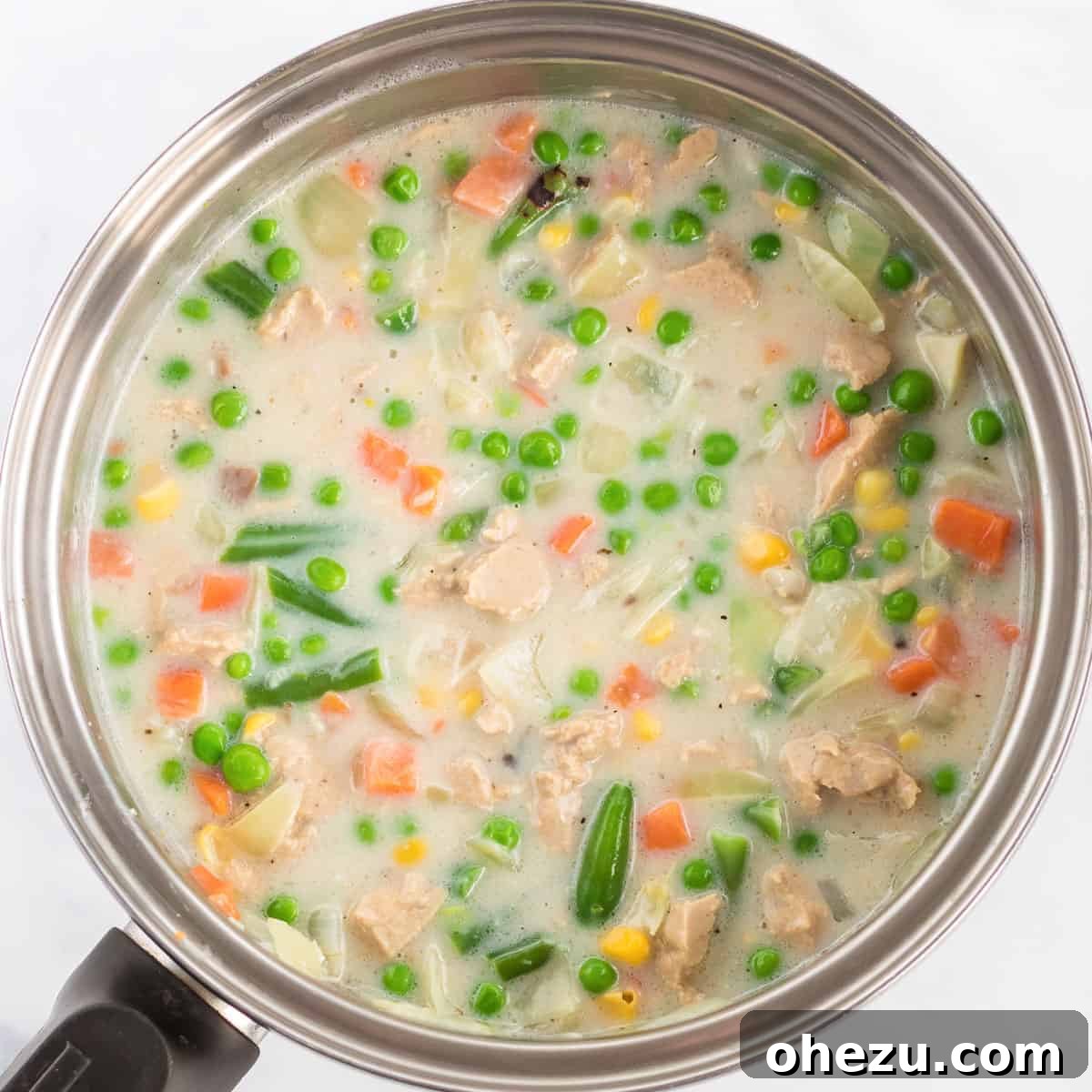Pouring fresh milk and adding mixed vegetables into a skillet with sautéed onions and garlic, beginning to form the creamy pot pie filling.