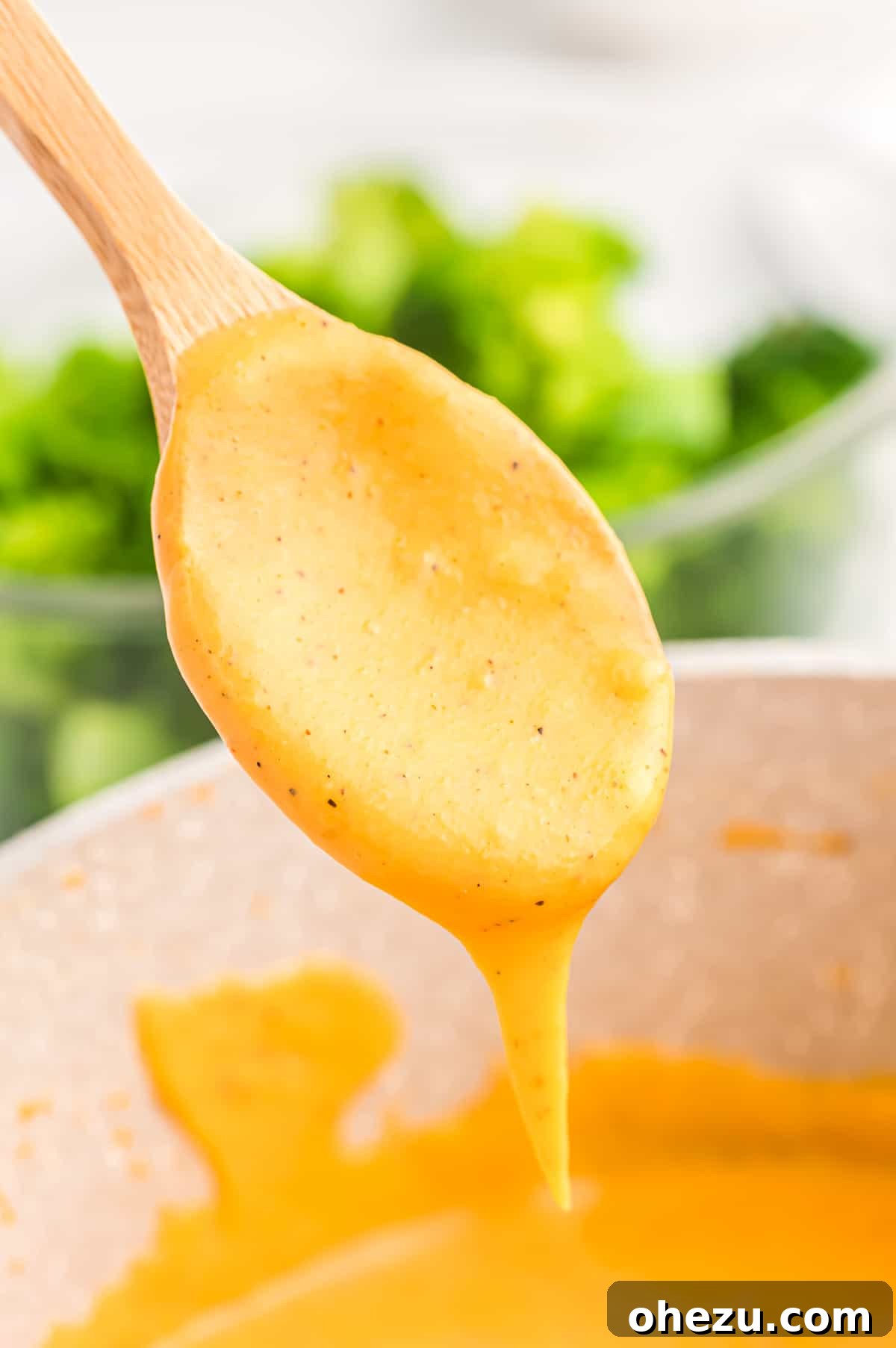 A spoon lifted from a saucepan, dripping with rich, golden cheddar cheese sauce, highlighting its creamy texture.