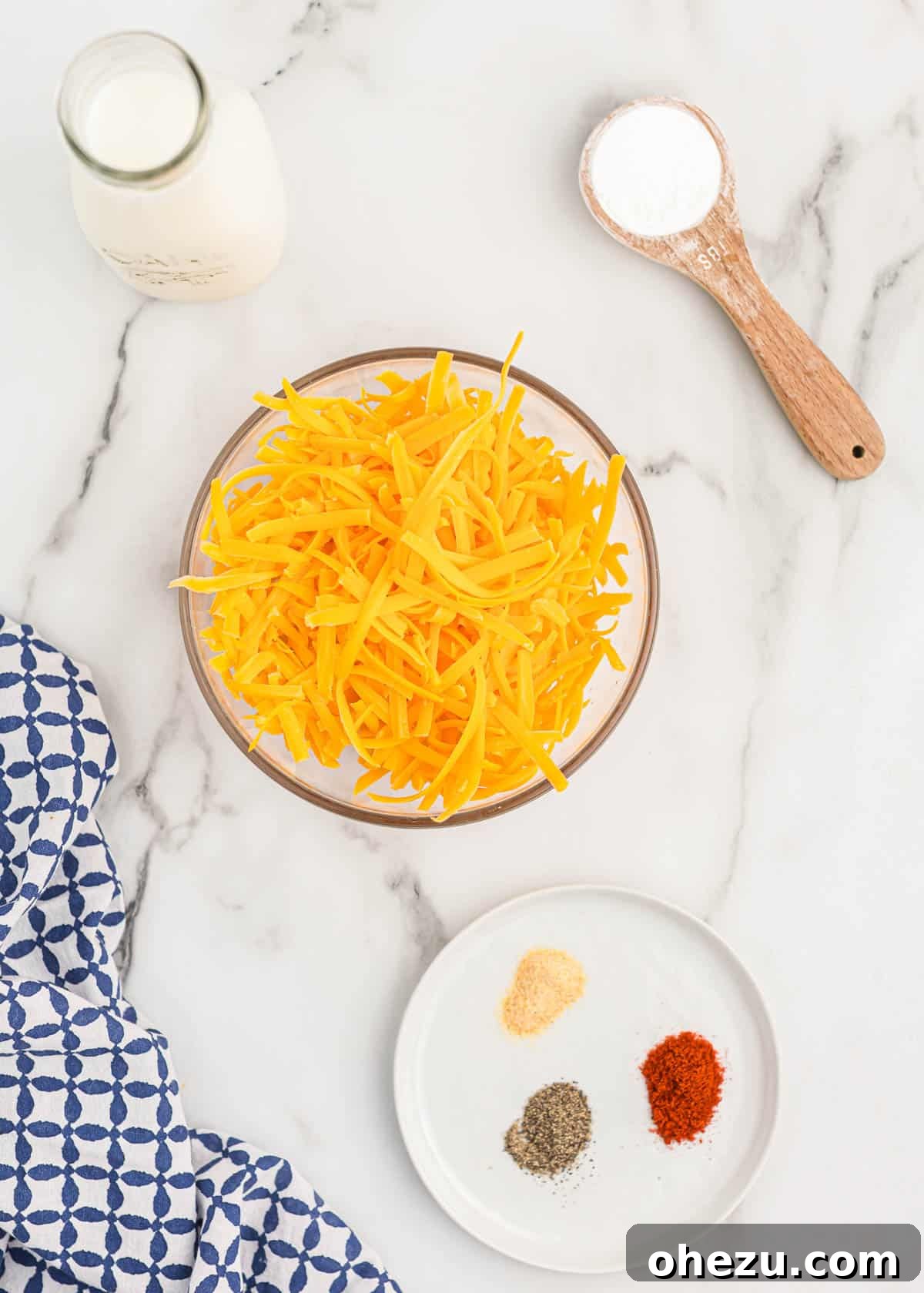 All the fresh ingredients laid out: a block of cheddar cheese, a bowl of grated cheese, milk, cornstarch, and small bowls of garlic powder, paprika, and black pepper.