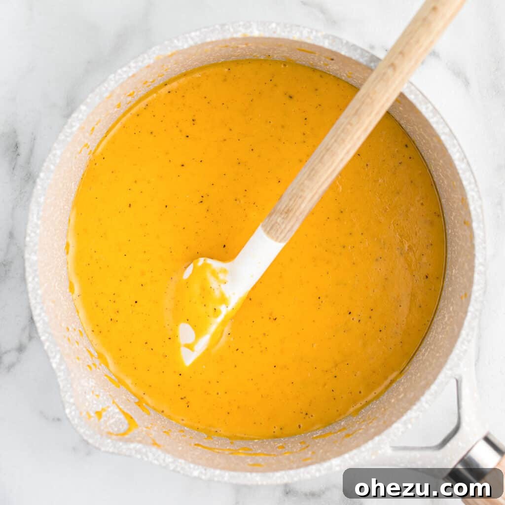 The cheese sauce for broccoli bubbling gently in a saucepan as it thickens, being stirred with a spoon.