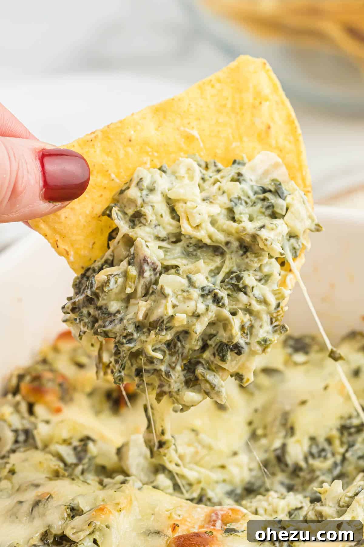A hand holding a tortilla chip, scooping up a generous portion of the warm, cheesy baked spinach artichoke dip, showing its inviting texture.