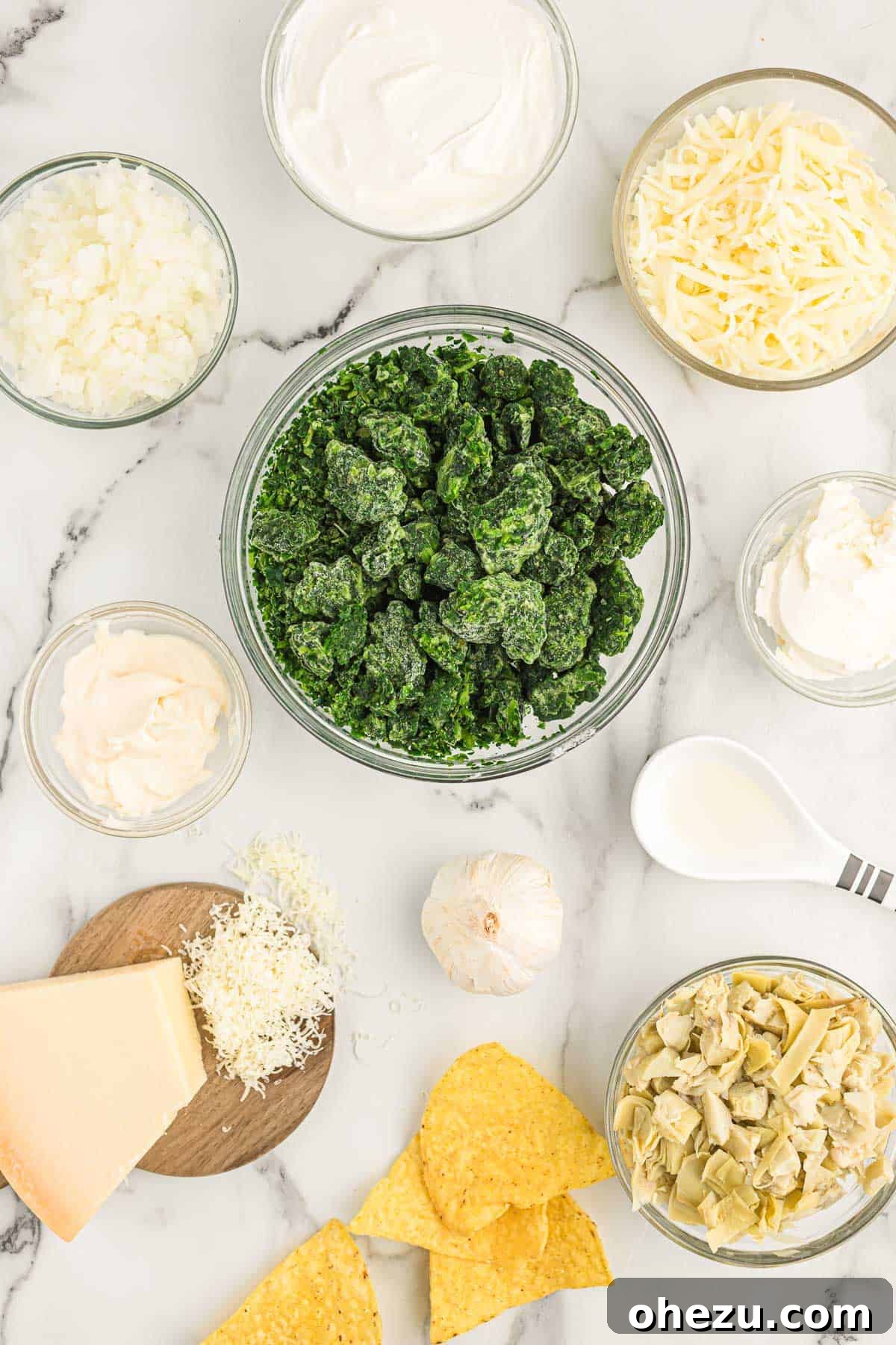 A flat lay photograph displaying all the fresh and packaged ingredients neatly arranged for making spinach artichoke dip, including a bowl of chopped artichokes, spinach, cheeses, and aromatics.