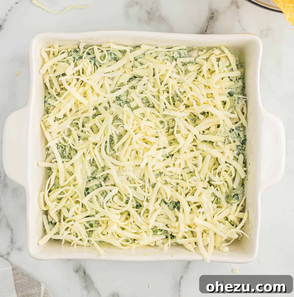 Spinach artichoke dip in a baking dish, generously topped with shredded mozzarella cheese, ready to be baked.