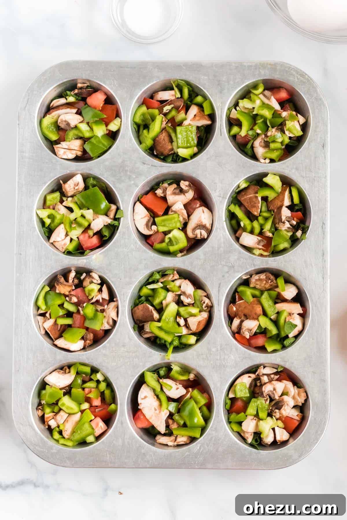 Finely chopped vegetables distributed evenly into a greased muffin tin, preparing for the egg mixture.