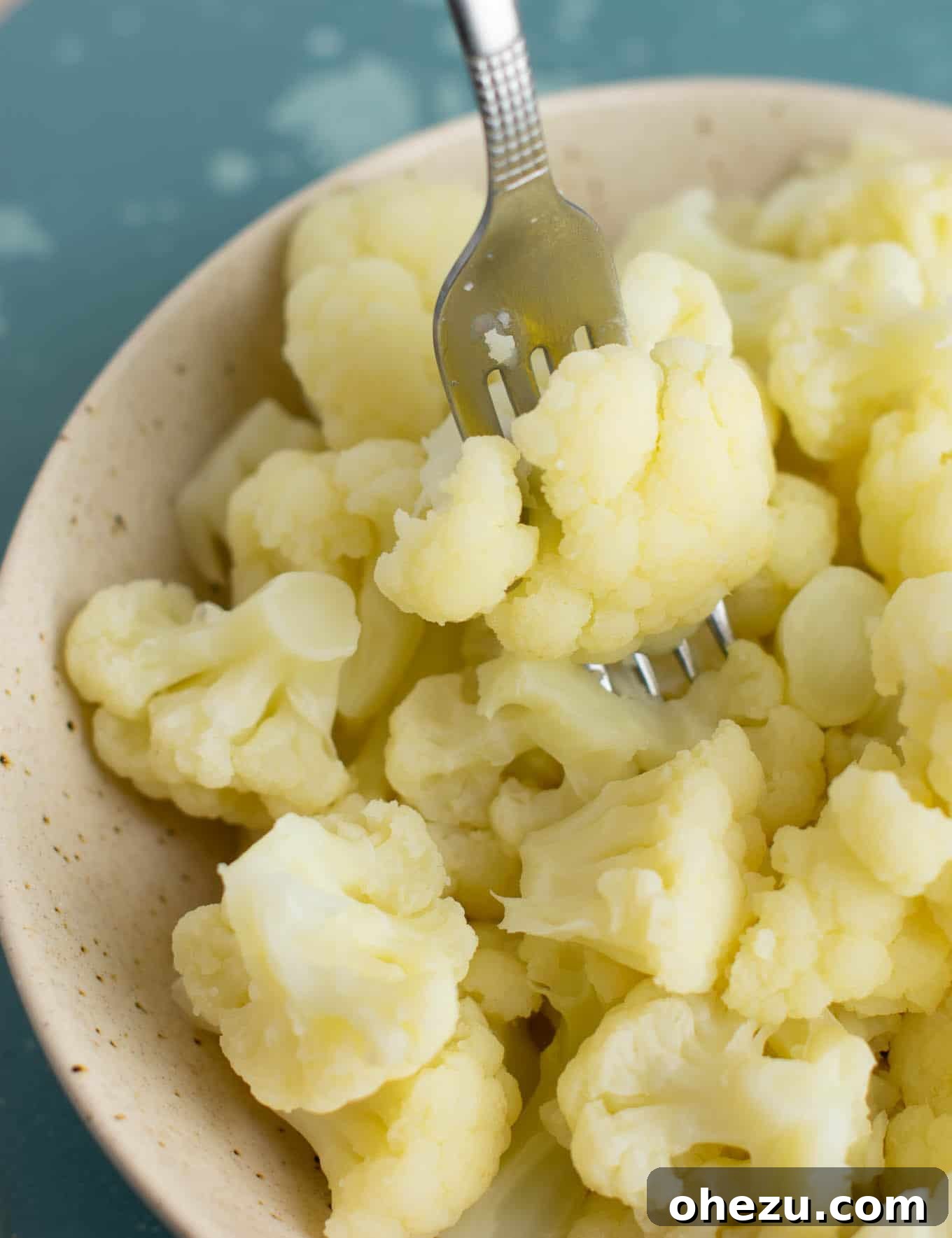 A fork lifting a tender piece of steamed cauliflower from a white bowl, showing its soft texture