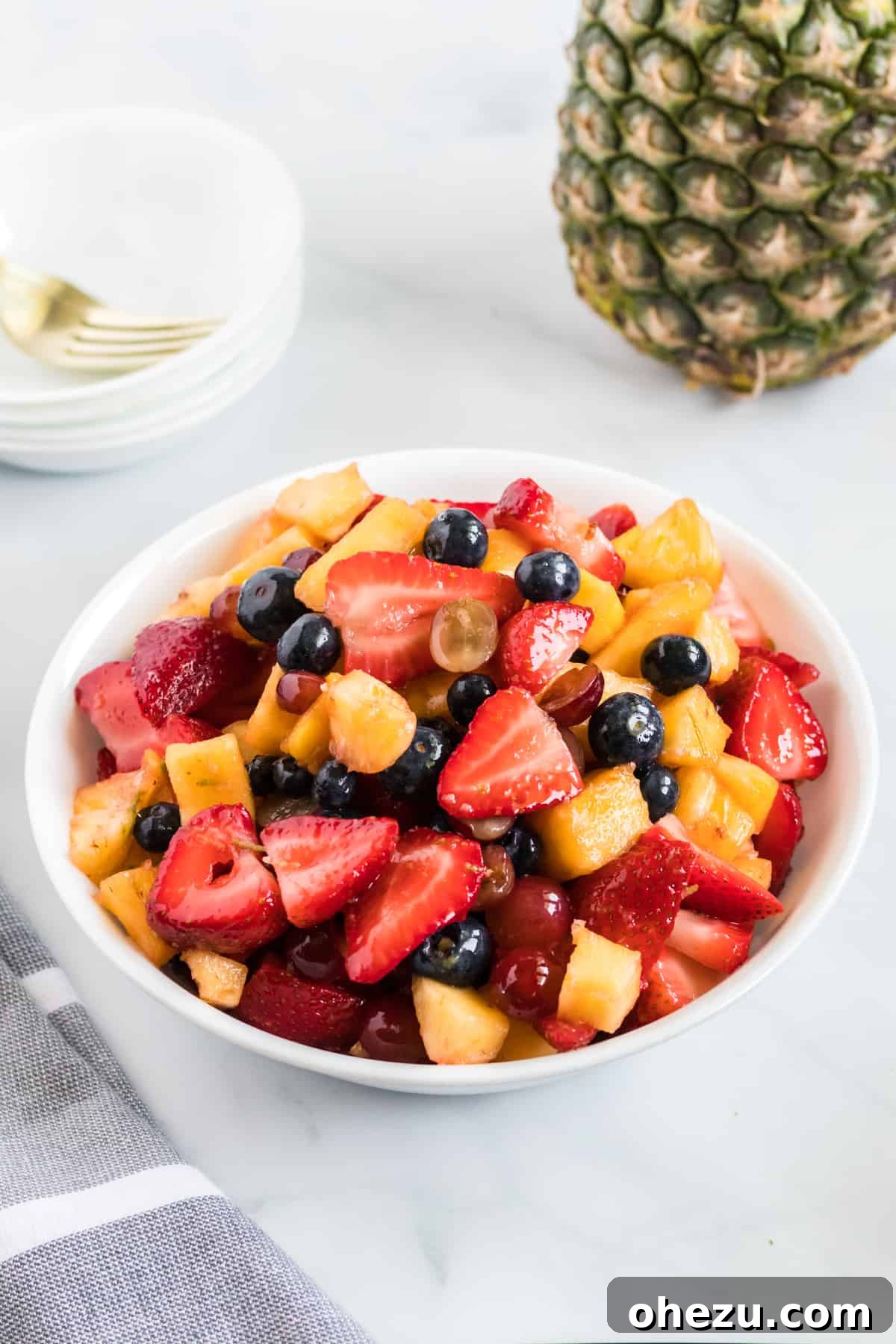 A small white serving bowl overflowing with vibrant, refreshing fruit salad, featuring pineapple, strawberries, blueberries, and grapes, glistening with honey-lime dressing.