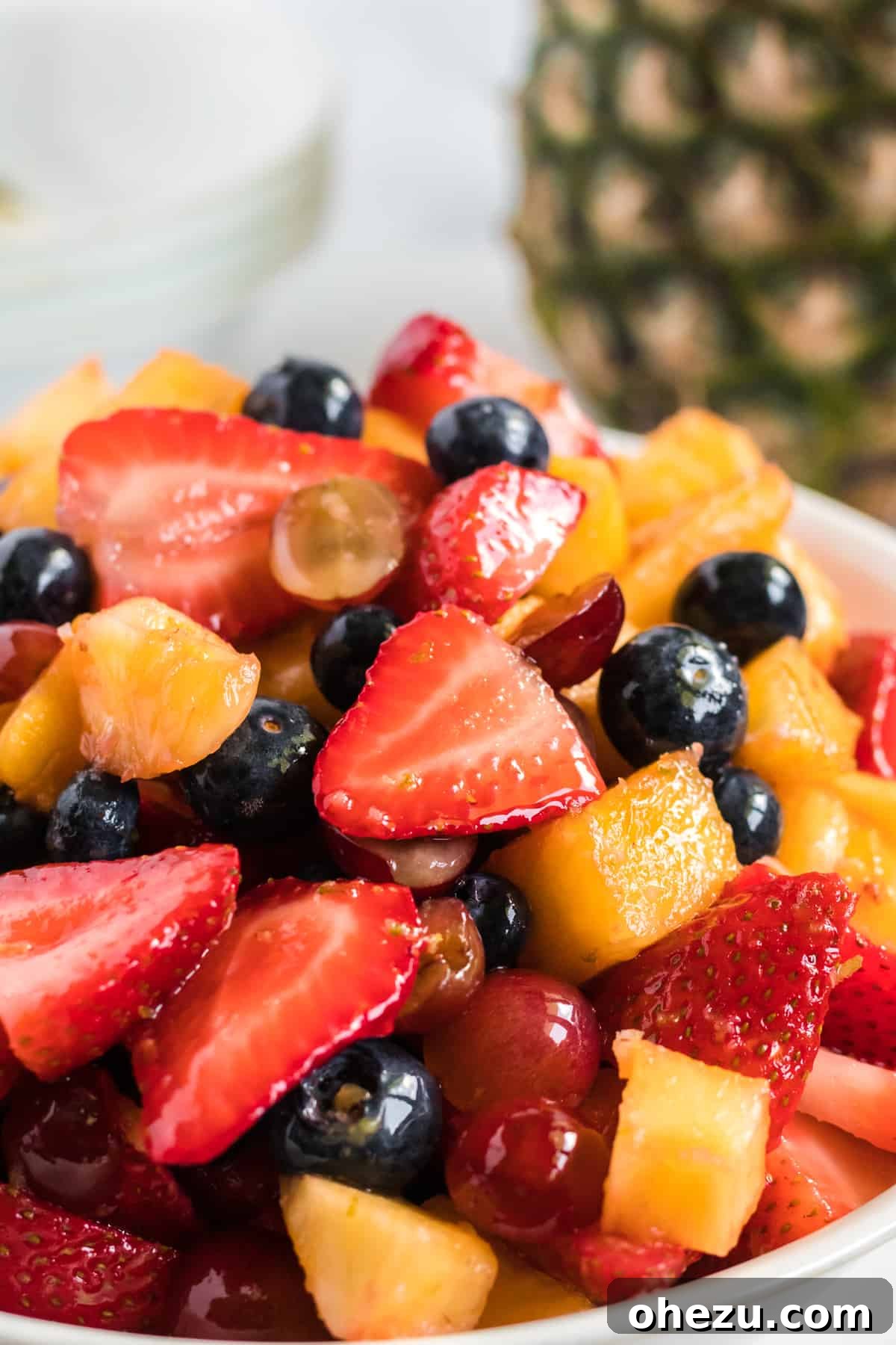 Close-up shot of the colorful fruit salad with a glossy honey-lime dressing, showing the texture of pineapple chunks, whole blueberries, sliced strawberries, and halved grapes, beautifully coated.