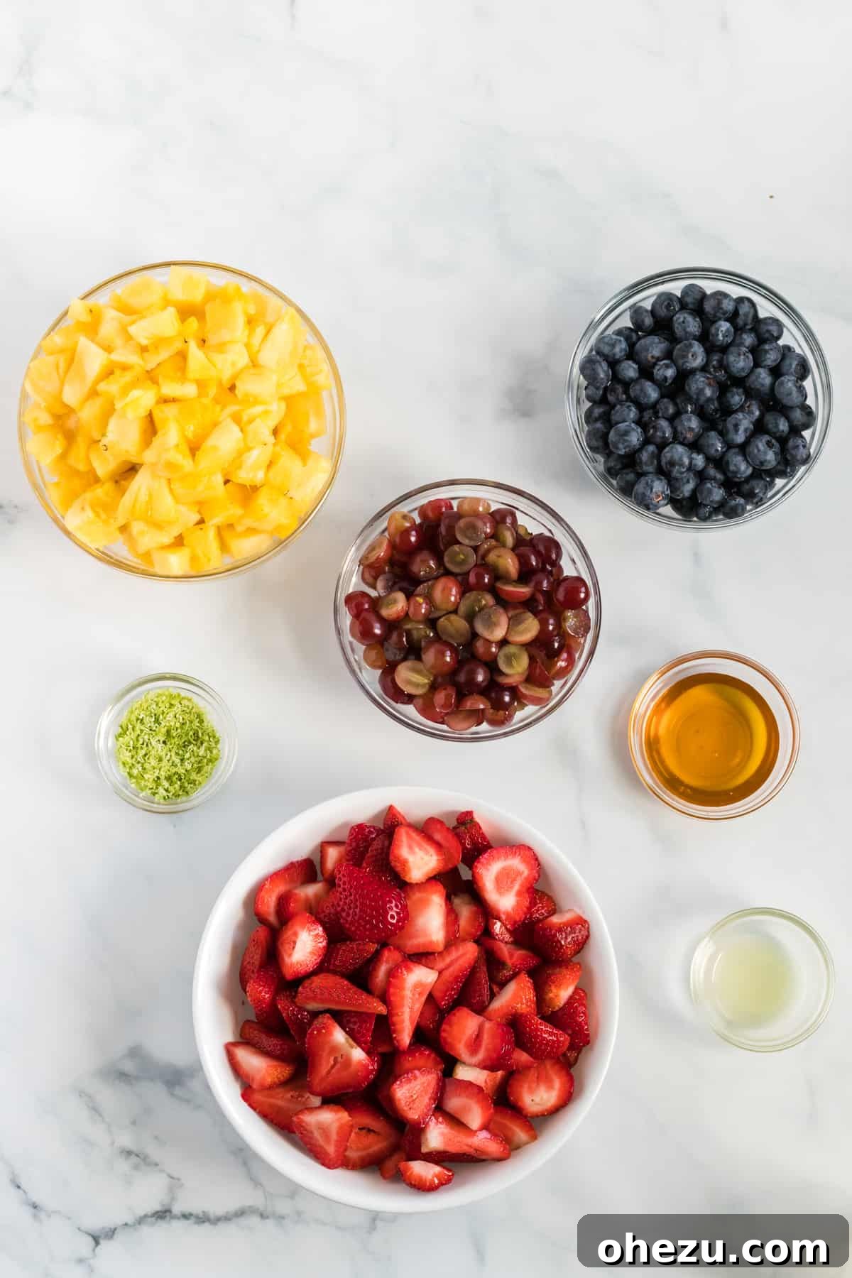 Separate glass bowls displaying the fresh ingredients for fruit salad: perfectly diced pineapple, vibrant sliced strawberries, plump fresh blueberries, halved grapes, golden honey, and a whole lime with its zest already grated, ready for mixing.