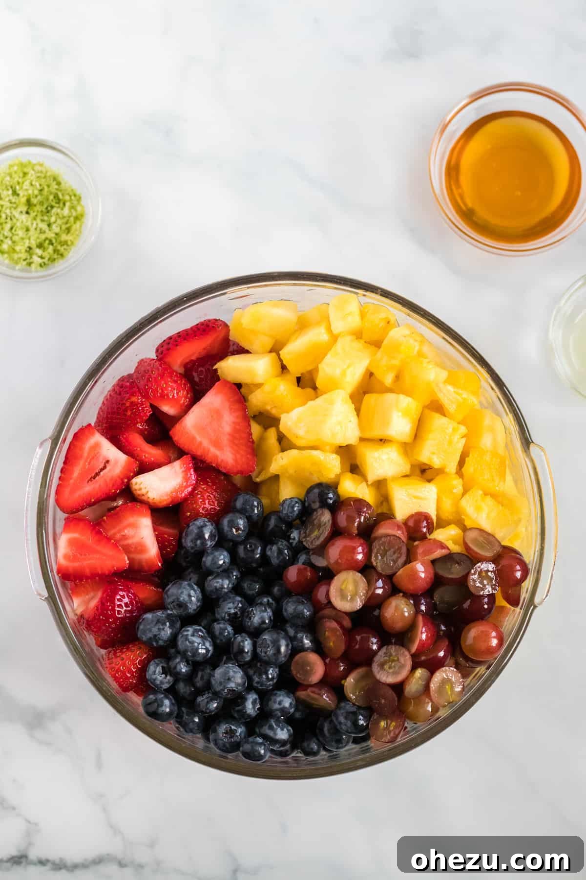 A large clear glass bowl filled with colorful, freshly prepared fruits: sliced strawberries, plump blueberries, halved grapes, and perfectly diced pineapple, awaiting the addition of the honey-lime dressing.