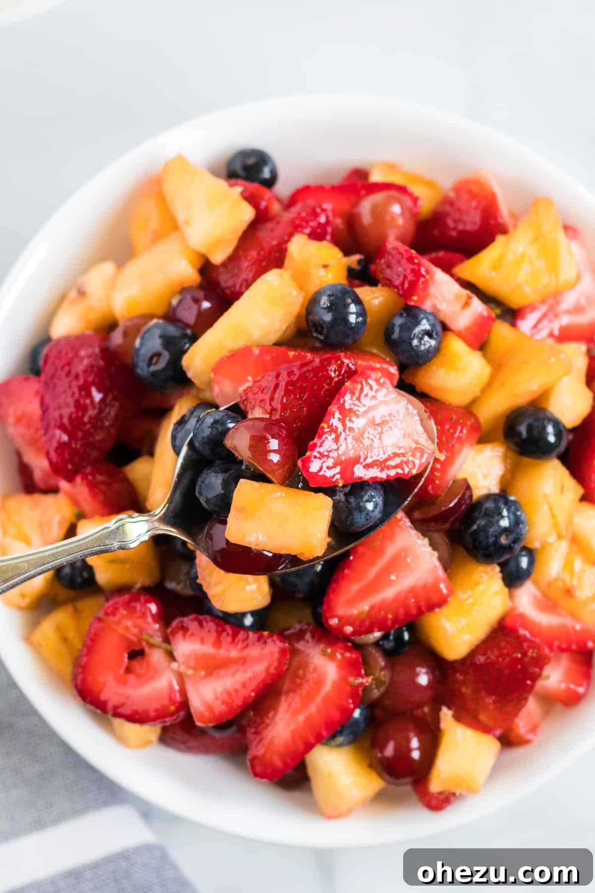 A spoon scooping a generous serving of colorful fruit salad from a large glass dish, highlighting the individual pieces of fruit glistening with dressing.