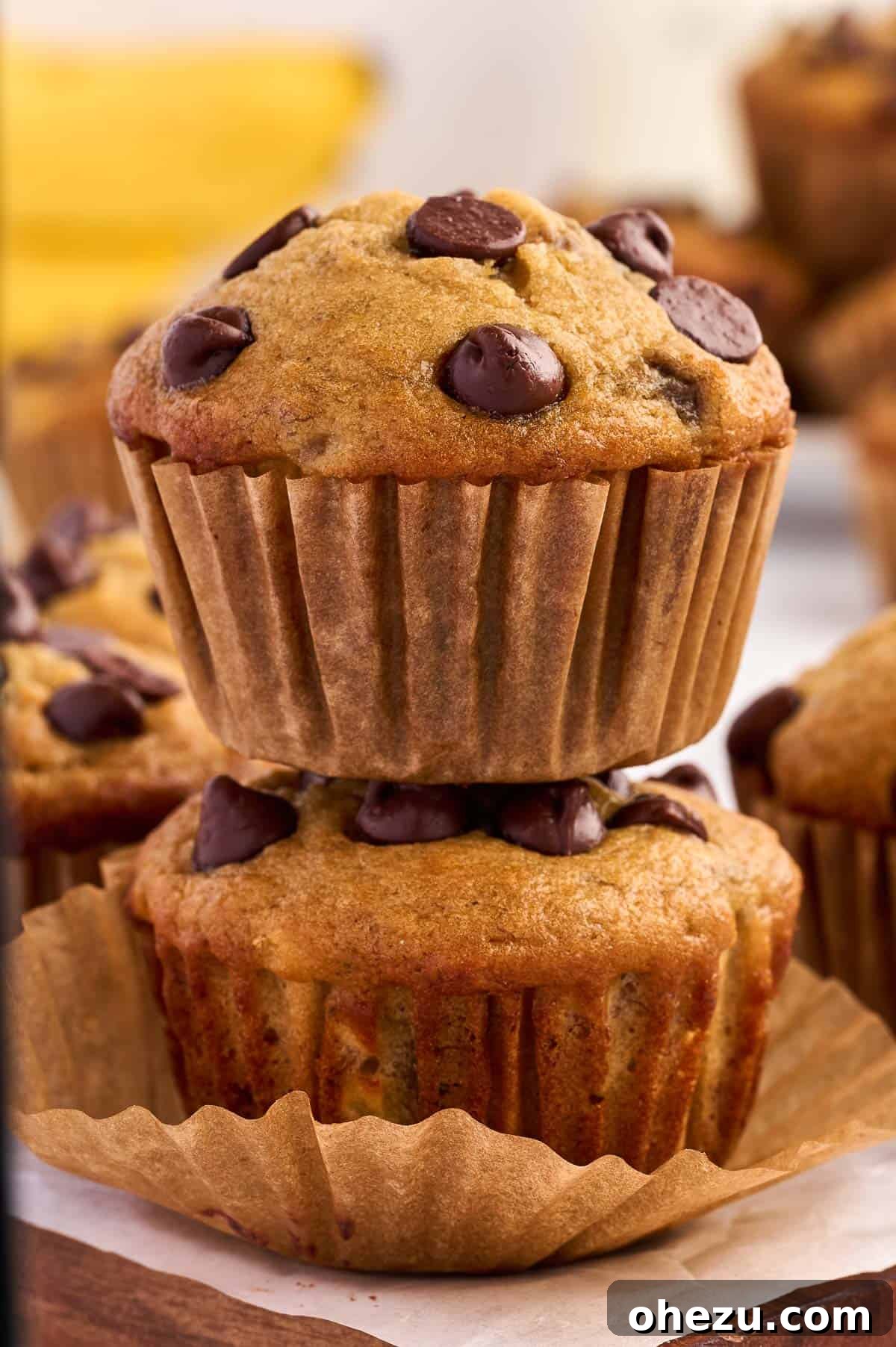 Moist Banana Greek Yogurt Muffins 16 A stack of three Greek yogurt banana muffins on a wooden board, with a blurred background of kitchen items.