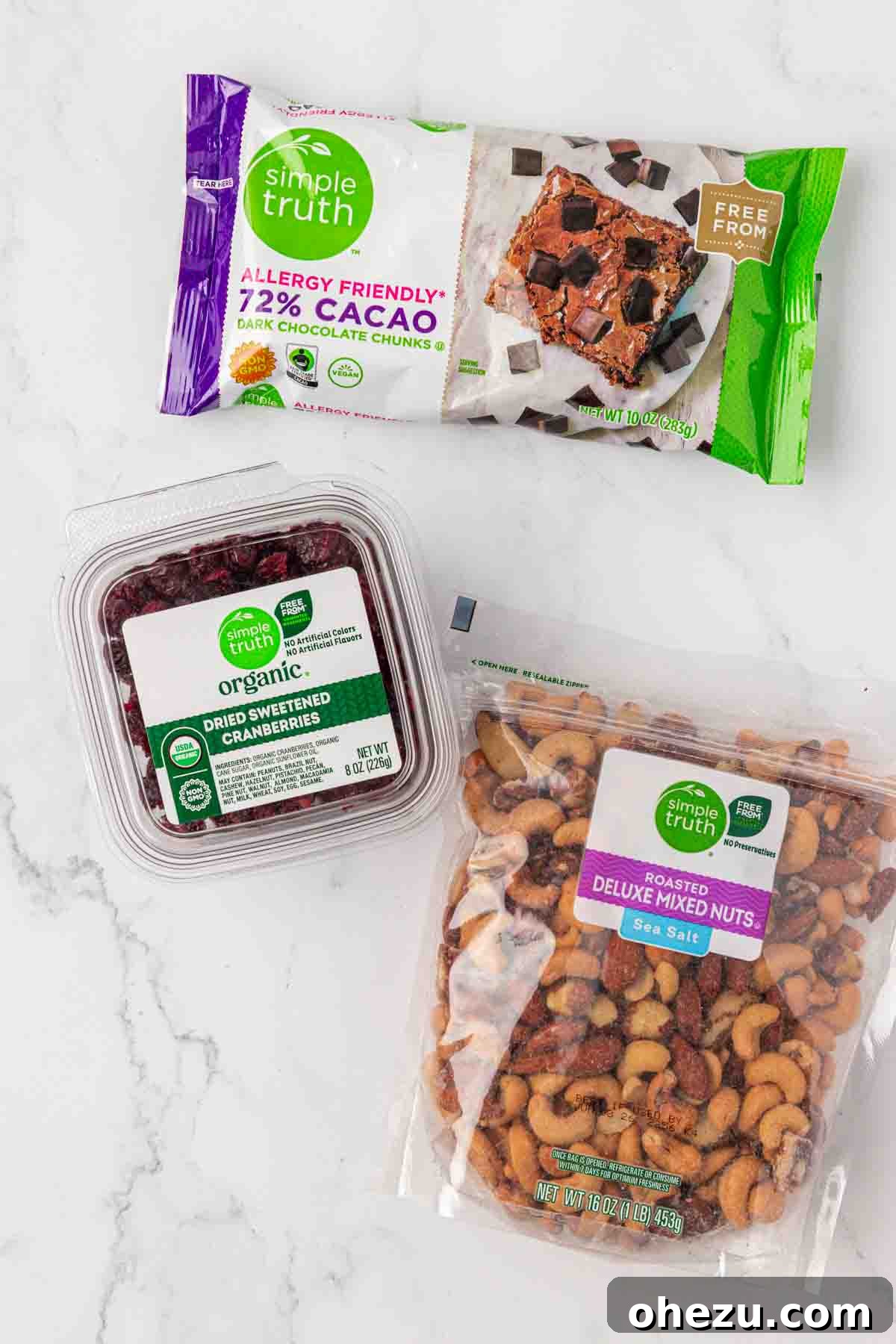 Three essential ingredients for homemade trail mix: a pile of rich dark chocolate chunks, a vibrant heap of dried sweetened cranberries, and a transparent bag of assorted roasted mixed nuts.