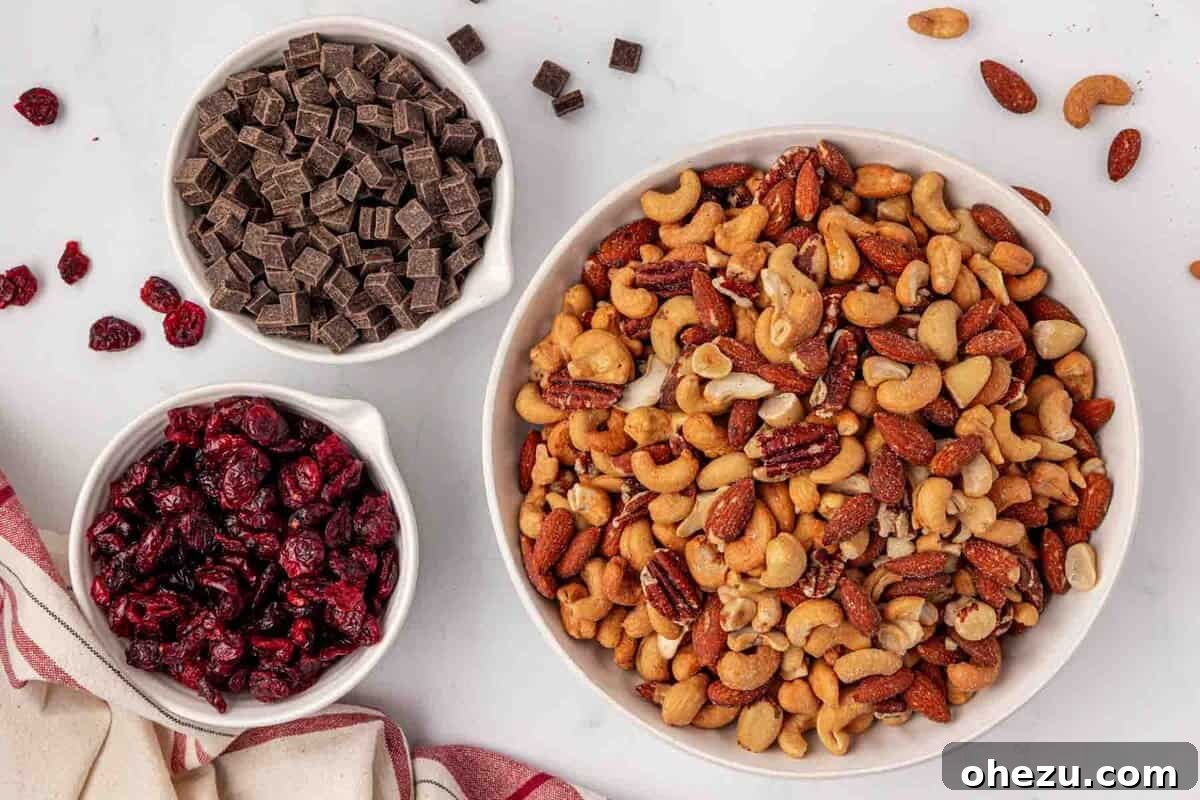 Three separate bowls containing the primary ingredients for homemade trail mix: one bowl with various roasted nuts, another with bright red dried cranberries, and a third with indulgent dark chocolate chunks, neatly arranged before mixing.