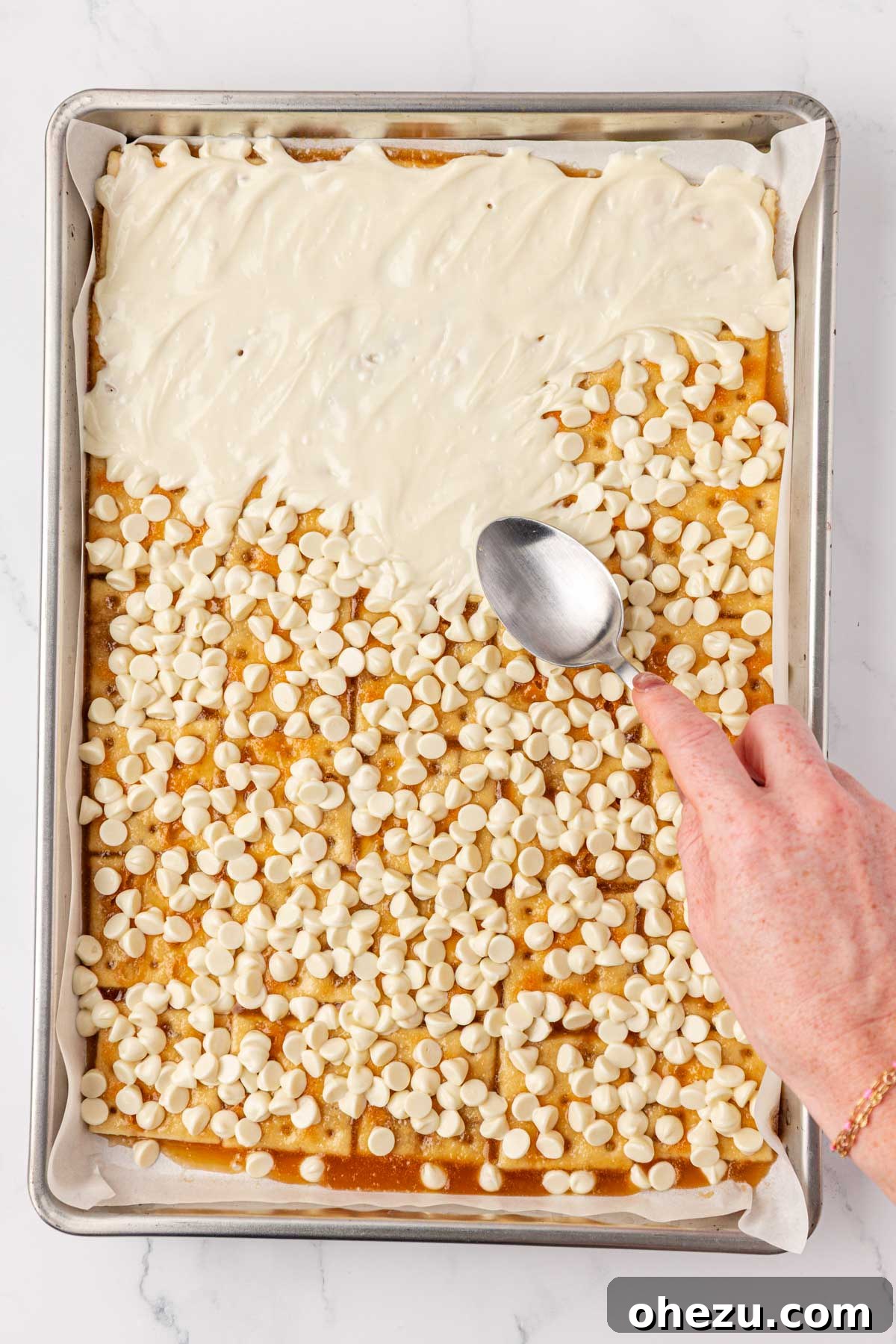 Using a spoon to gently spread the melting white chocolate chips over the toffee-covered saltine crackers.