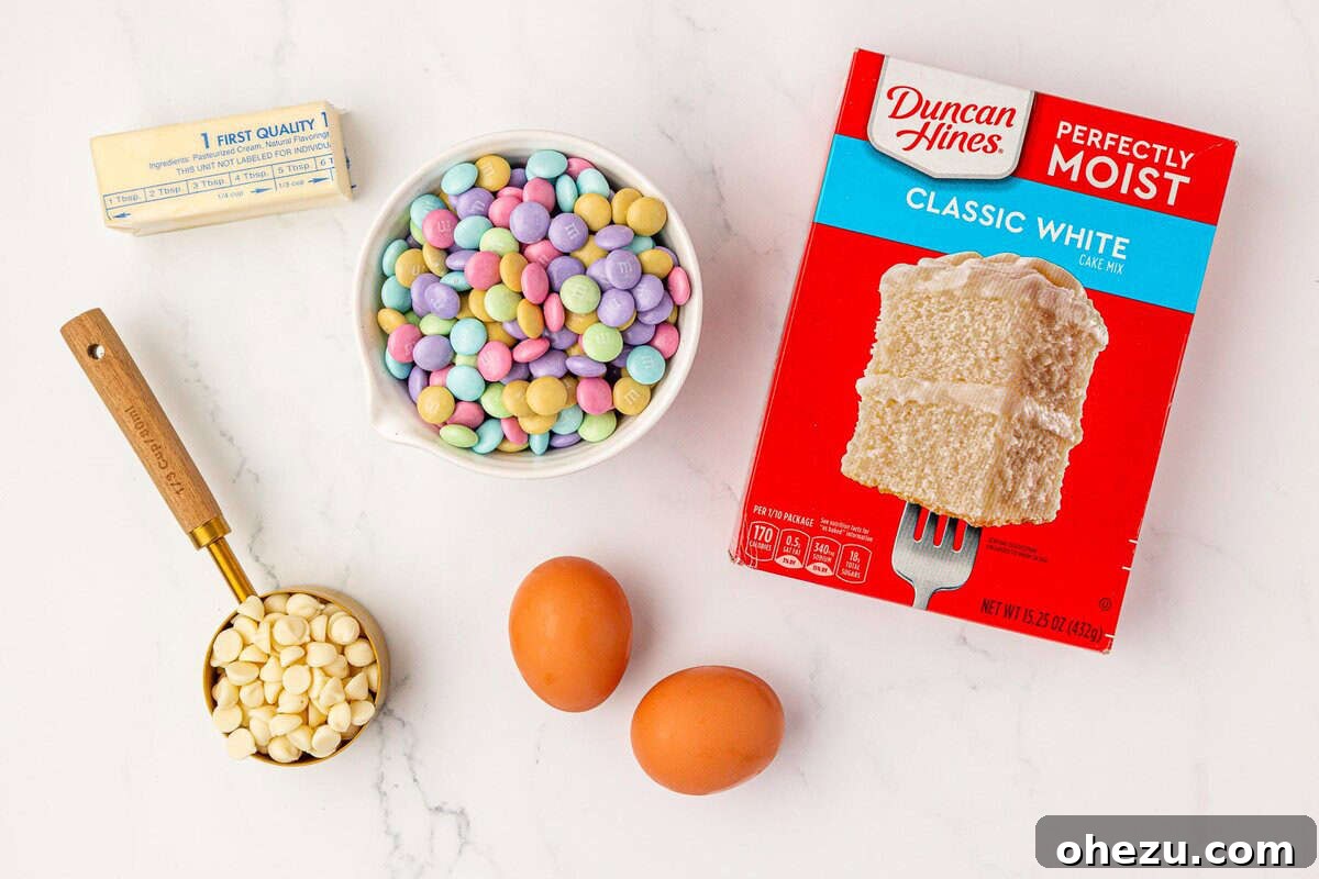 A flat lay of all 5 ingredients: melted butter, white chocolate chips, Easter M&M's, eggs, and vanilla cake mix ready for baking.