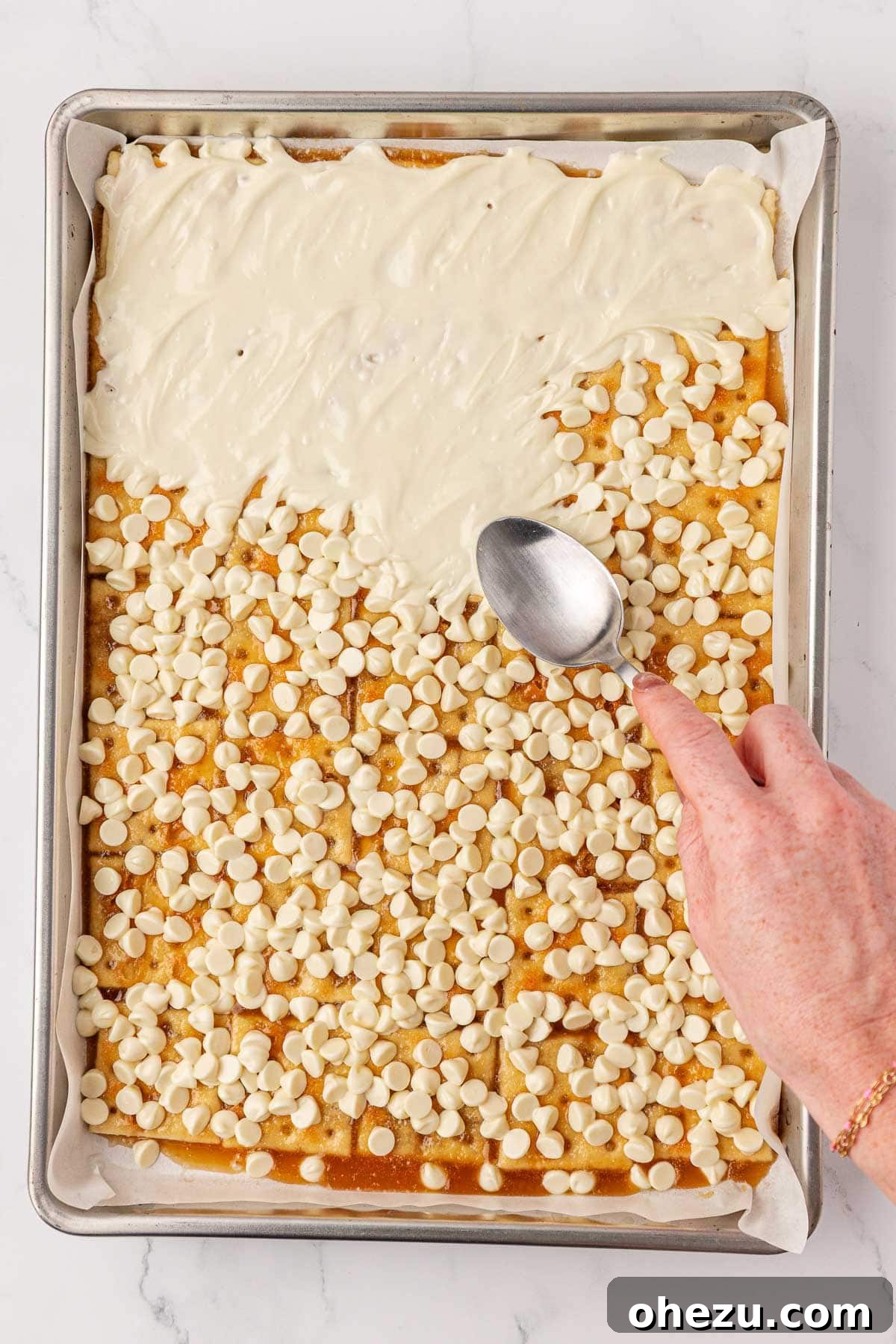 Skillfully spreading the melted white chocolate chips across the surface of the toffee crackers using the back of a spoon